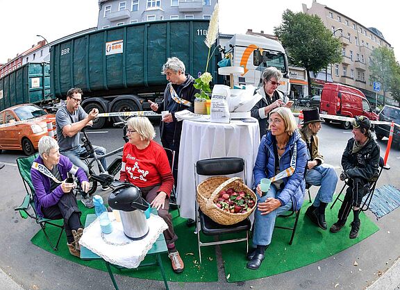 Auf dem Parking Day 2019 in Berlin-Tempelhof haben Menschen einen Tisch mit Kaffee und Obst aufgestellt und unterhalten sich in entspannter Atmosphäre.