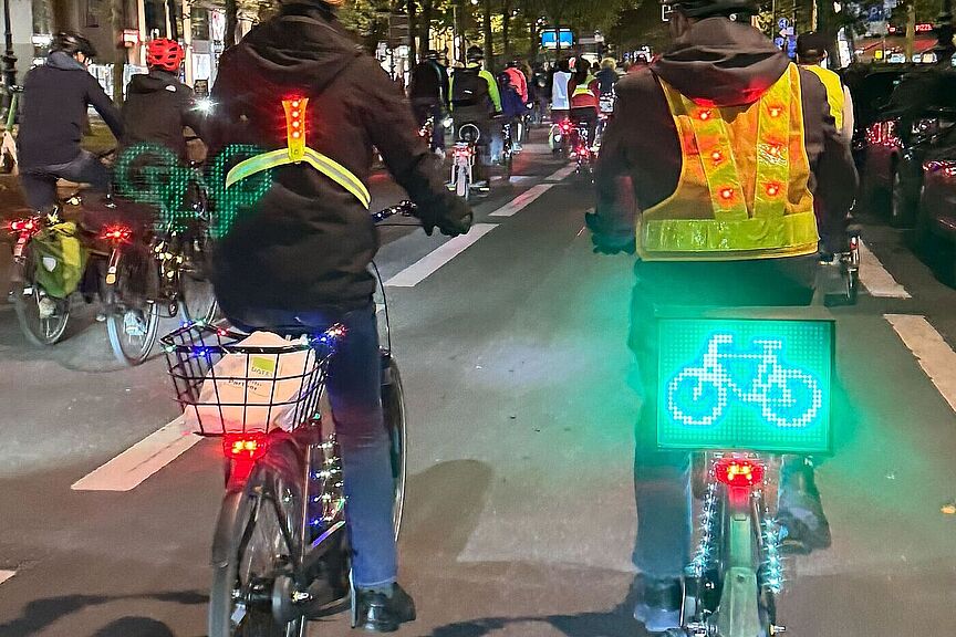 Beleuchteter Fahrradprotest ADFC-Lichterfahrt Berlin, 10. Oktober 2025: Mehrere Menschen fahren auf beleuchteten Fahrrädern durch eine Fußgängerzone. An den Fahrrädern leuchten rote und grüne LED-Lichter, ein Fahrrad trägt ein leuchtend grünes Display mit Fahrrad-Symbol. Die Radfahrenden tragen fluoreszierende Sicherheitswesten. Im Hintergrund sind weitere beleuchtete Fahrräder zu sehen.